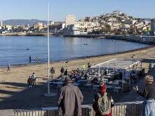 Plage animée avec des personnes, bateaux et bâtiments en arrière-plan.